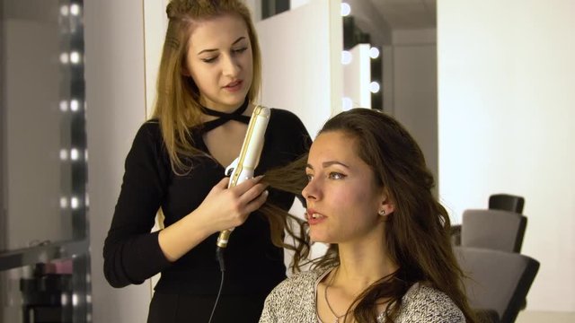 Young beautiful girl in beauty salon. Hairdresser-stylist makes the girl's hair. The hairdresser curls the hair with a styler and stacks them.