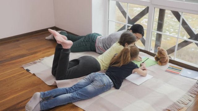 Mother with two daughters draws pictures relaxing on floor near the window