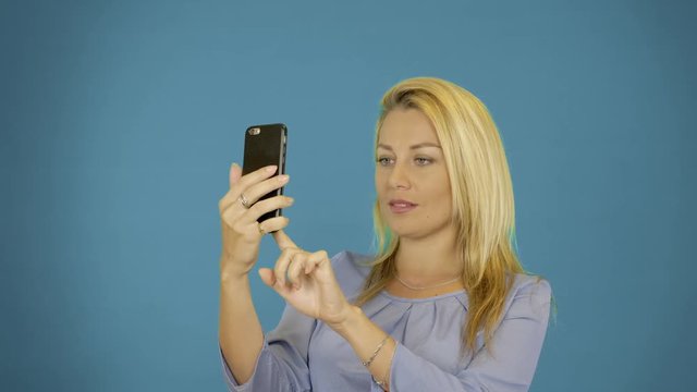 Young caucasian woman take photo on smartphone. Solid blue background. Female portrait waist up. Pretty blonde smiling.