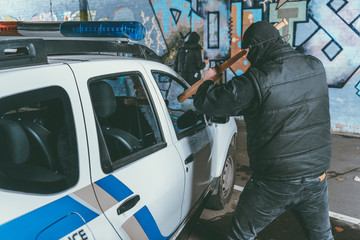 Fototapeta premium vandal crashing police car with baseball bat while another man painting graffiti on wall