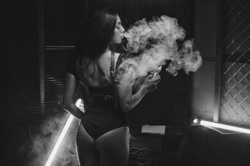Young beautiful brunette woman smoking, vaping e-cigarette with smoke on black background.