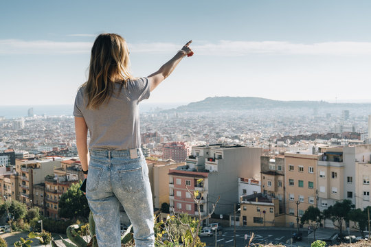 Rear view of young woman wearing in stripped t-shirt standing on high point and looking at cityscape, pointing at something. Summer sunny day, rear view, bird's eye view of city, cityscape, horizon.