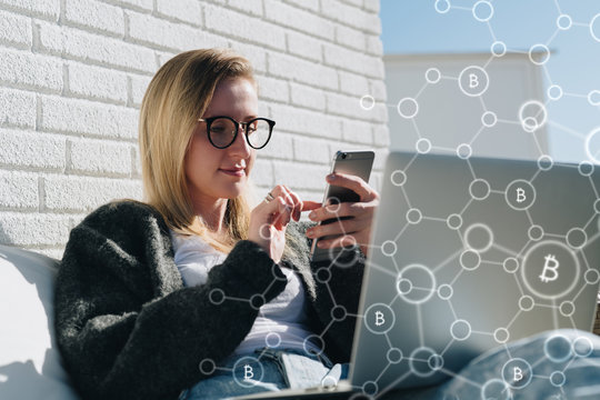 Young business woman in glasses sits at laptop and uses smartphone. In foreground infographics, bitcoin icons. Girl working, learning online. Online marketing, education, e-learning.Business planning.