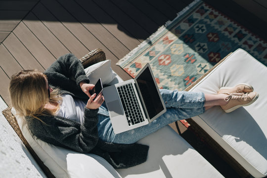 Sunny day. View from above. Young businesswoman is sitting on white couch on terrace, using laptop and smartphone. Girl working, blogging, learning online. Distance work. Online marketing, education.
