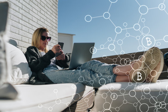 Young business woman in sunglasses sits and uses smartphone, laptop. In foreground infographics,bitcoin icons. Girl working, learning online. Online marketing, education, e-learning.Business planning.