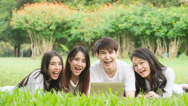 Group of Asian students laying on grass outdoor park, with laptop books and happy smile for friendship and education concept.