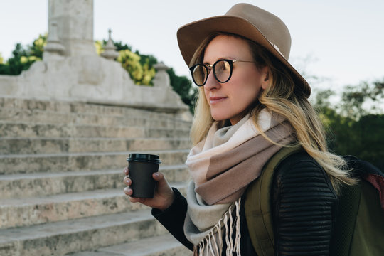 Sunny autumn day. Young attractive woman travels in hat, eyeglasses and with backpack stands on city street, drinks coffee.Hipster girl is looking for road, sights.Vacation,adventure, trip.
