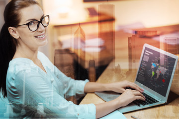 Busy bee. Cheerful young woman in eyeglasses sitting at the table and posing while creating a presentation for her project on laptop