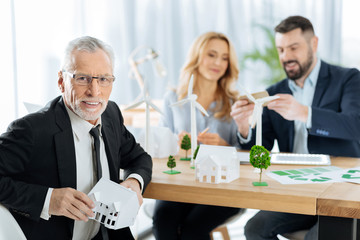 Successful project. Pleasant cheerful aged man sitting at the table with a lovely little house in his hands while his friendly colleagues sitting not far from him