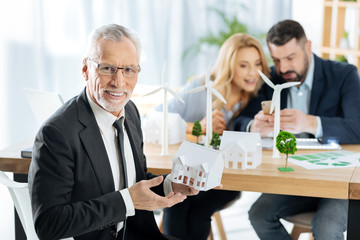 Important building. Smart excited aged man showing a lovely little while house while his skilled creative colleagues sitting at the table and discussing the construction of windmill turbines