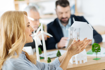 Lovely idea. Happy experienced clever engineer sitting next to her colleagues and admiring a lovely little model of an eco friendly house while holding it in her hands