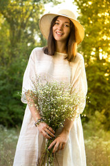 Beautiful sunny smiling girl / woman in white dress and straw hat with bouquet of wildflowers - 185234904
