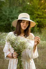 Beautiful sunny smiling girl / woman in white dress and straw hat with bouquet of wildflowers - 185234921
