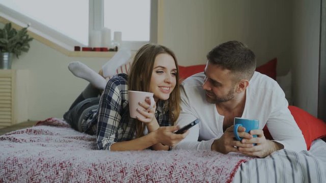 Smiling couple watching TV while lying in bed and drinking coffee at home in the morning