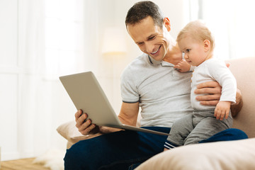 Fatherhood. Friendly positive young father sitting on a sofa and looking at the screen of a laptop while spending time with his cute little son