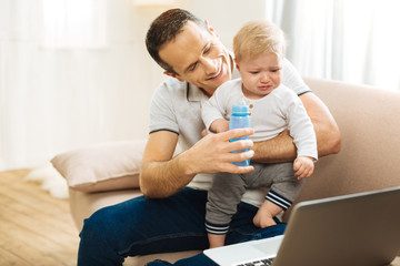 Stop crying. Kind attentive cheerful father feeling responsible while sitting on a soft sofa and giving a bottle of water to his upset crying child