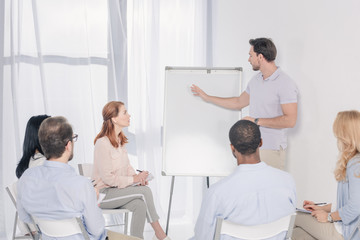 Obraz premium psychotherapist pointing at blank whiteboard and multiethnic group sitting on chairs during therapy