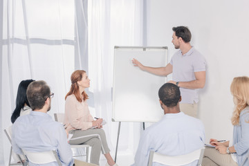 psychotherapist pointing at blank whiteboard and multiethnic group sitting on chairs during therapy