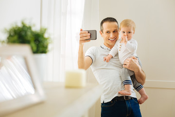 Delighted father. Cheerful emotional young man standing near the big window and smiling while holding a baby and taking photos with him