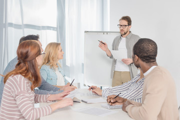 middle aged man in in eyeglasses with papers standing at whiteboard and having conversation with his team