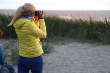 blonde Frau in blau-gelbem Anzug von hinten mit Kamera fotografiert einen Sonnenuntergang über der...