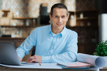 Show your emotions. Delighted man keeping smile on his face and putting elbows on the table while looking straight at camera