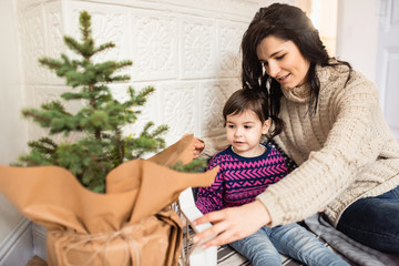 Horizontal portrait of beautiful Caucasian mom and daughter decorate the Christmas tree indoors with wood star, candles. Portrait loving mother and toddler girl together.