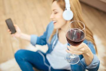 Drinking wine. Overjoyed blond smiling long-haired woman sitting on the carpet with her eyes closed and with headphones and holding her phone and a glass of red wine while listening to music