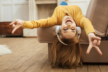 Being energetic. Pretty overjoyed young blond woman laughing and listening to music and wearing headphones while lying on the sofa and wearing a yellow sweater