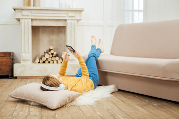 Weekend. Young blond slim woman listening to music and wearing headphones and holding her phone while lying on the floor and a cushion under her head and a fireplace in the background