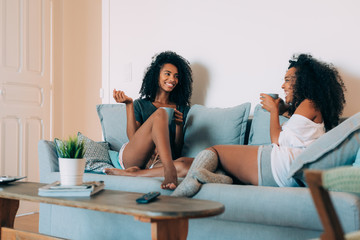 Happy young two black women sitting in the couch drinking coffee .