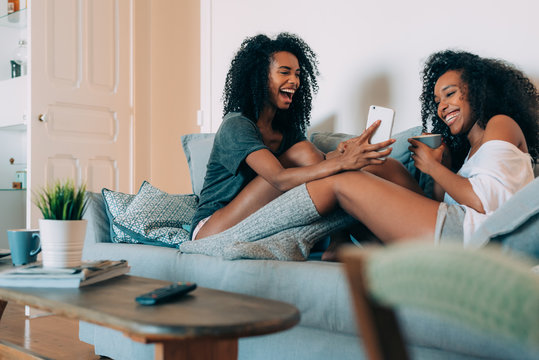 Happy young two black women sitting in the couch looking at the mobile phone and drinking coffee .