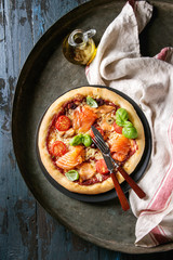 Traditional pizza with smoked salmon, cheese, tomatoes and basil served on black plate with kitchen towel and olive oil over old dark metal background. Top view with space. Rustic style