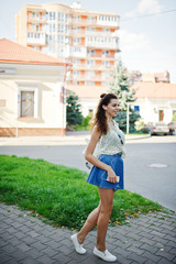 Fototapeta premium Curly stylish girl wear on blue jeans skirt, blouse and listening music from headphones phone. Portrait on streets of city.