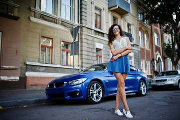 Curly stylish girl wear on blue jeans skirt, blouse and glasses posed near blue car at street of city.