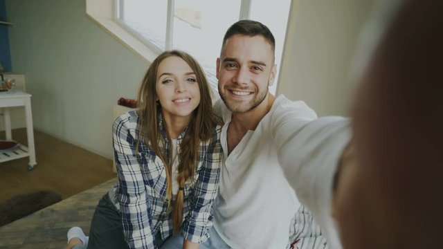 POV of Young smiling and beautiful couple take selfie portrait on smartphone camera and kiss while sitting on bed in the morning