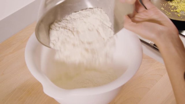 A woman pours flour from a metallic bowl into a plastic one in a kitchen - closeup from above