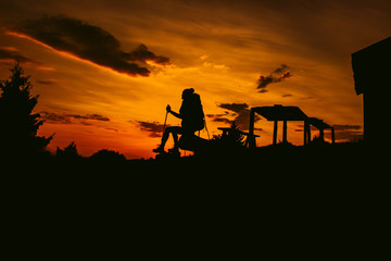 Silhouette hiker woman sit on the bench, with backpack and trekking pole, sunset orange sky on the background. Rest during a hike