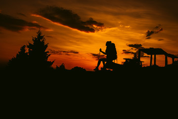 Silhouette hiker woman sit on the bench, with backpack and trekking pole, sunset orange sky on the background. Rest during a hike