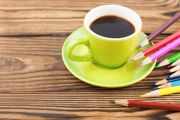 Lot of colorful sharpened pencils scattered on old wooden rustic brown table beside ceramic cup full black coffee on green saucer