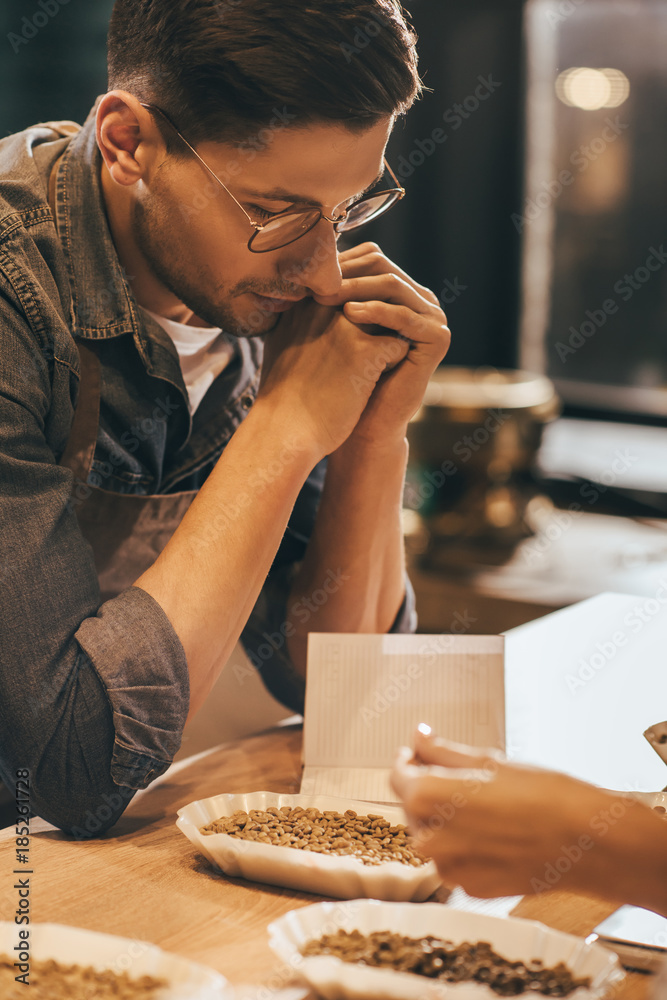 Wall mural side view of pensive coffee shop worker looking at coffee beans in bowl on wooden tabletop