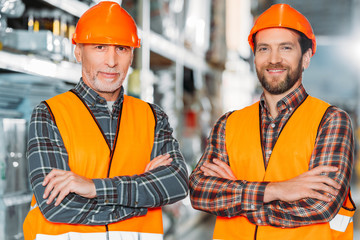 two male workers in safety vests and helmets with crossed arms in storehouse