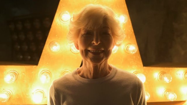 Happy retirement. Cheerful nice ederly woman smiling while standing against illumination