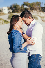 Pregnant young couple interacting facing each other, standing outdoors, beach scene, embracing, with casual wear.