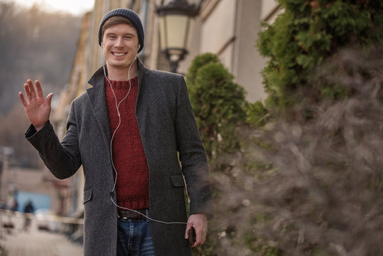 Half-length portrait of charming male walking in the city with phone and headset. He is expressing openness and friendliness