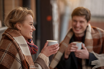 Laughing man and woman enjoying coffee with blankets. Focus on lady while boy is looking at her expressing affection