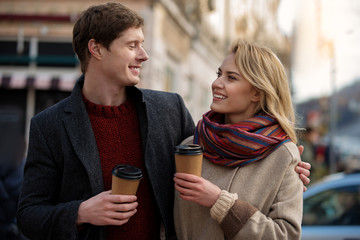 Waist up portrait of amorous man and woman standing together in the street. Male is tenderly hugging female while lady is smiling