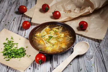 vegetarian soup of fresh greens, vegetables and pasta in a brown plate, chopped green onion on brown baking paper and cherry tomatoes on a light wooden table