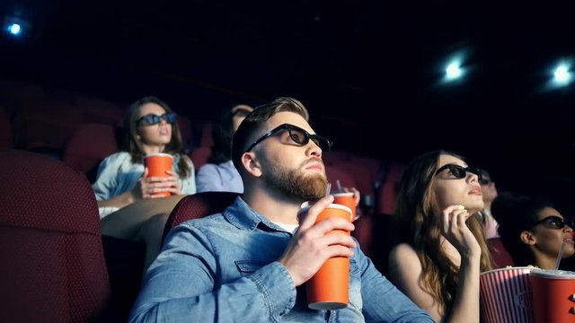 Young modern men and women in 3d glasses watching film and eating popcorn in cinema