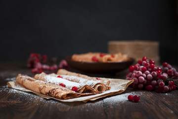 sweet fresh pancakes with a pile and pancakes on baking paper, rolled with tubules, viburnum berries on a dark wooden table on a dark gray background